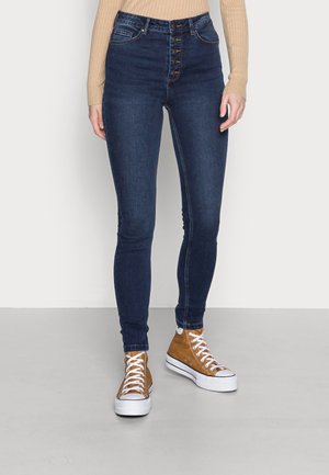Onderlichaam met donkerblauwe high-waisted skinny jeans met vijf knopen aan de voorkant en bruine hoge sneakers met witte veters en zolen.