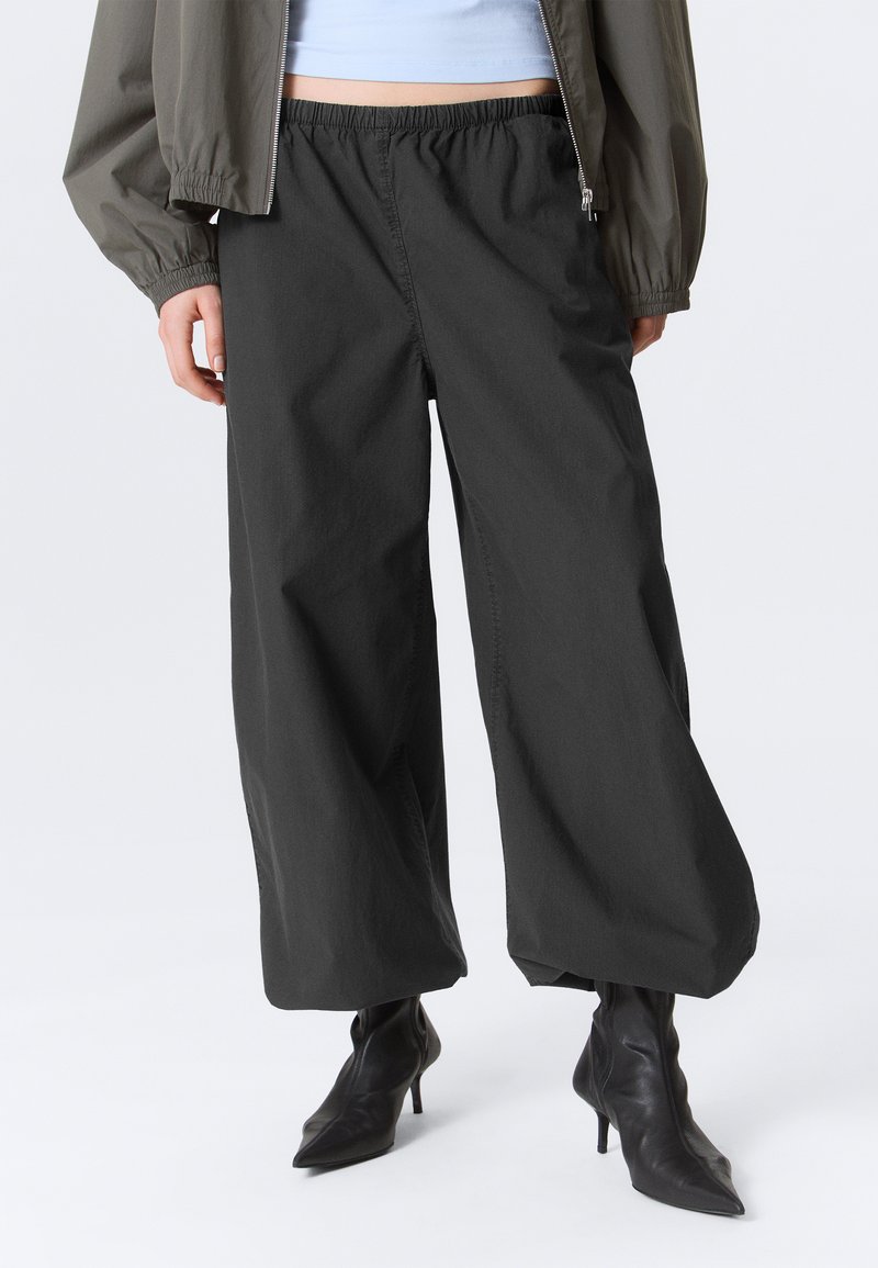 Personne portant un pantalon noir à jambes larges, des bottines à talons pointues noires, un haut blanc court et une veste verte olive zippée.