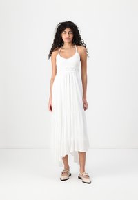 White maxi dress with a crocheted bodice, spaghetti straps, and tiered hemline; worn with light-colored slip-on shoes.
