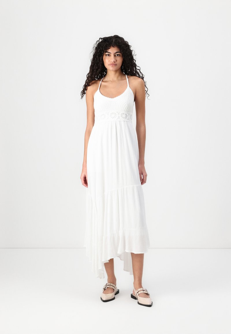 White maxi dress with a crocheted bodice, spaghetti straps, and tiered hemline; worn with light-colored slip-on shoes.