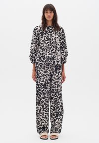 Femeie în picioare, purtând un set coordonat format dintr-un top lejer negru și bej cu imprimeu abstract și pantaloni largi, completat cu sandale bej, pe un fundal alb.