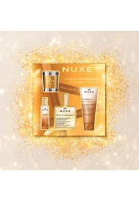Caja de regalo dorada que contiene productos de NUXE: una botella de vidrio, un tubo de crema y un tarro pequeño, todos con etiquetas elegantes y diseños sofisticados.