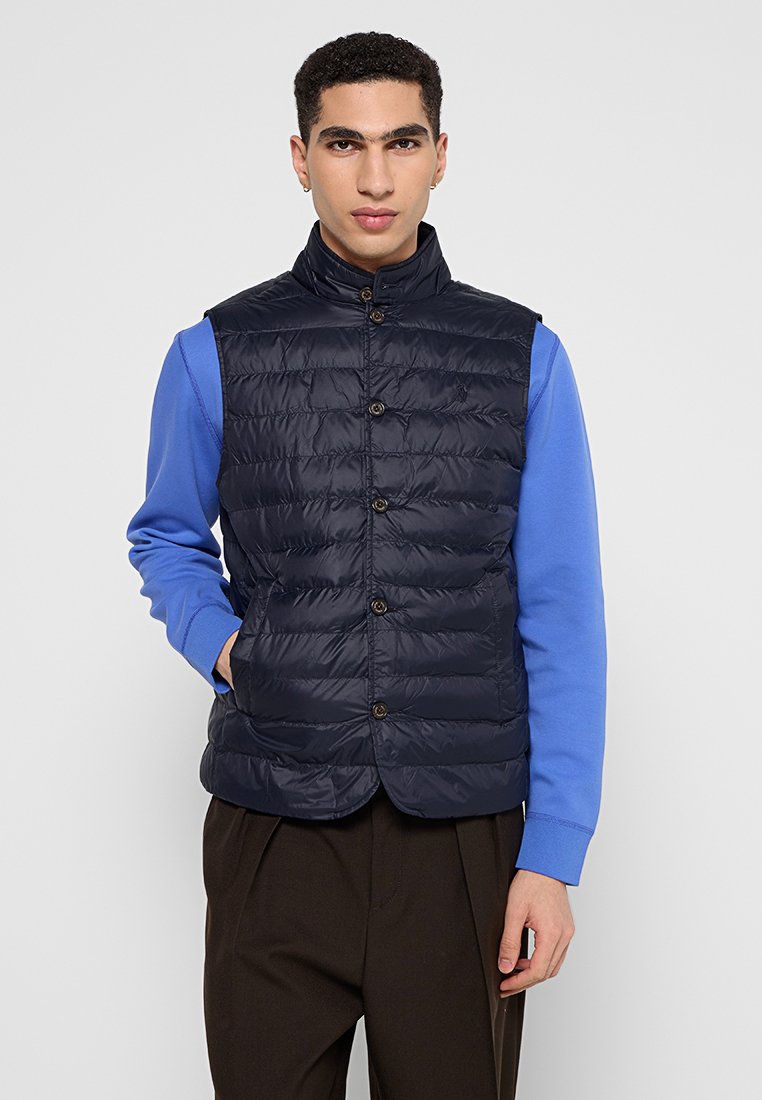 Granatowa pikowana kamizelka z wysokim kołnierzem, zapinana na guziki z przodu, o gładkiej, lekkiej fakturze, zestawiona z niebieską koszulą z długim rękawem.