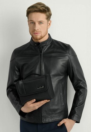 Młody mężczyzna z krótkimi jasnobrązowymi włosami i brodą, ubrany w czarną skórzaną kurtkę, trzyma czarną kosmetyczkę na zamek.