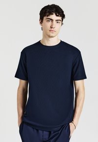 Giovane uomo con capelli corti e ricci di colore scuro, che indossa una camicia corta con maniche e tessuto strutturato di colore blu navy e pantaloni abbinati, su uno sfondo semplice.