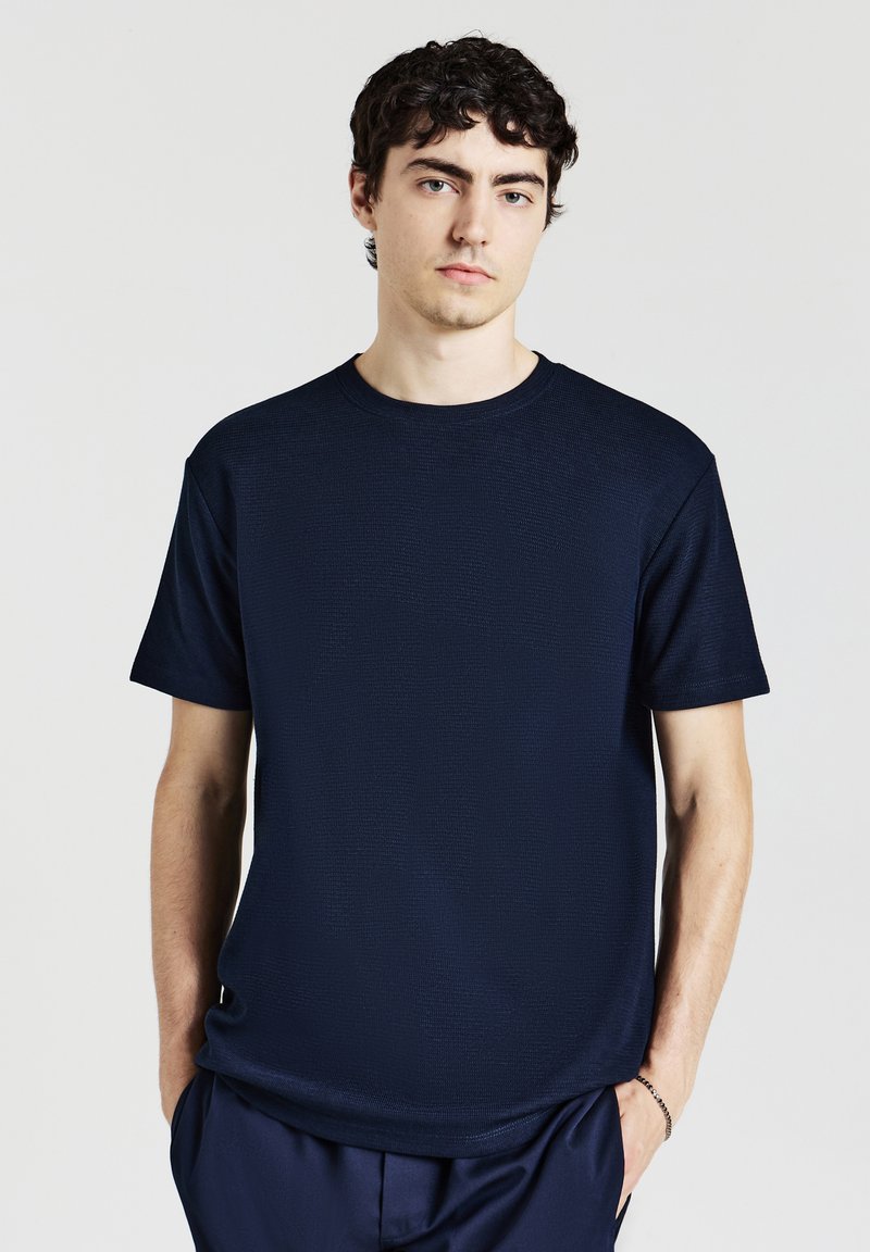 Giovane uomo con capelli corti e ricci di colore scuro, che indossa una camicia corta con maniche e tessuto strutturato di colore blu navy e pantaloni abbinati, su uno sfondo semplice.