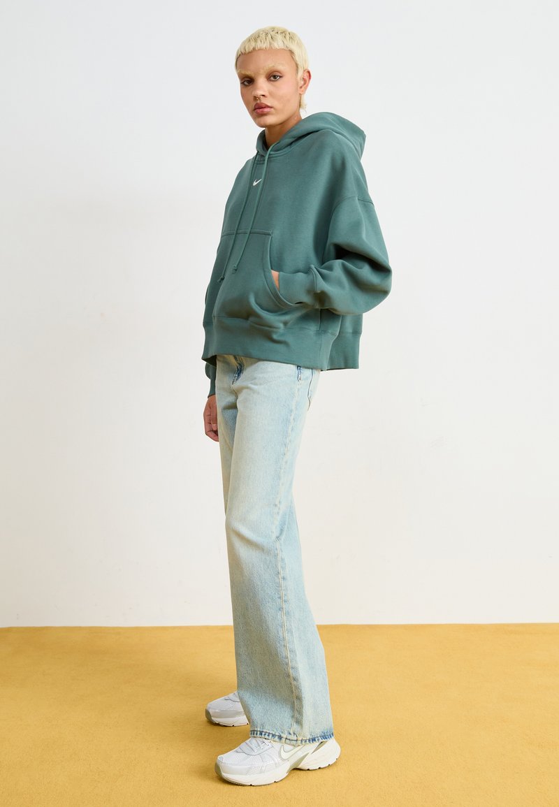Sweatshirt surdimensionné teal avec une poche avant et une capuche à cordon, associé à un jean droit délavé et des baskets blanches.