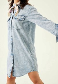 Ljusblå denimklänningsskjorta med långa ärmar, två bröstfickor och rundad nederkant. Har en lätt sliten textur och knappstängning fram.