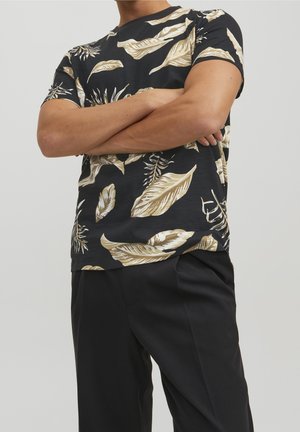 Black short-sleeve t-shirt featuring a floral design in beige, with leaves and plants. Paired with black trousers, showcasing a relaxed fit.