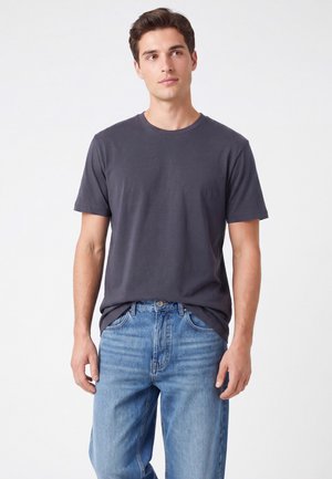 Giovane uomo con una maglietta grigio scuro semplice e jeans blu, in piedi, che guarda leggermente di lato su uno sfondo bianco.