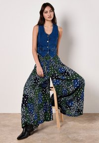 Chaleco de mezclilla con cierre de botón, combinado con pantalones de diseño floral de pierna ancha en azul y verde. Modelo lleva botas de tobillo negras.