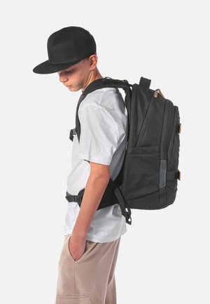 Black backpack with a textured fabric, padded straps, and buckles. Worn on a person in a white shirt and tan pants, facing sideways.