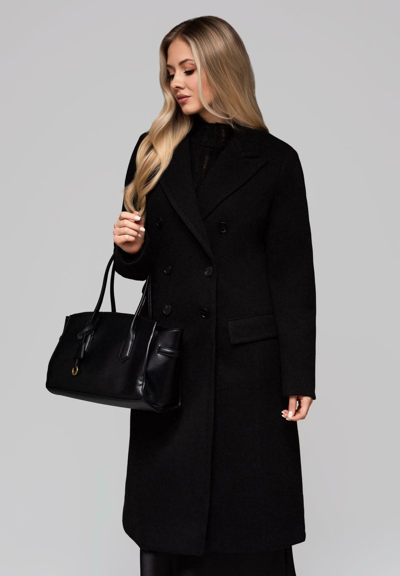 Donna con lunghi capelli biondi che indossa un cappotto nero doppiopetto, mentre tiene una borsa in pelle nera contro uno sfondo grigio uniforme.