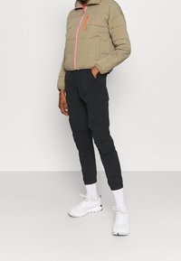 Giacca puffer color khaki con zip rosa, abbinata a pantaloni cargo neri. Sneakers bianche e calzini alla caviglia completano l'outfit.