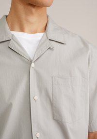 Camisa de manga corta de color gris claro con cuello, dos botones y un bolsillo en el pecho. Textura de tejido suave y detalles de diseño mínimos.