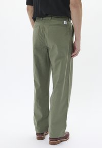 Pantaloni ușori, de culoare verde măsliniu, cu o croială lejeră, două buzunare la spate și un etichetă de marcă vizibilă pe talie. Se poartă încălțăminte maro.