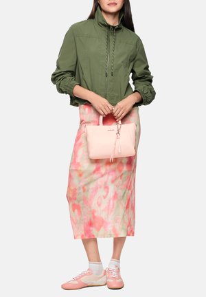 Frau trägt grüne Jacke mit Reißverschluss, rosa-beige gemusterten Midirock, rosa Sneakers, hält eine hellrosa Handtasche vor weißem Hintergrund.