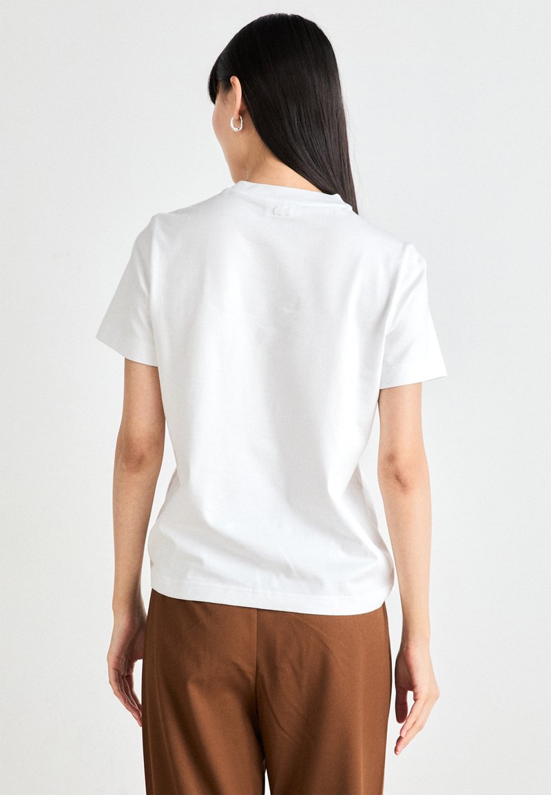 T-shirt bianca in cotone con scollo rotondo, maniche corte e design della schiena senza cuciture. Abbinata a pantaloni marroni. Texture semplice.