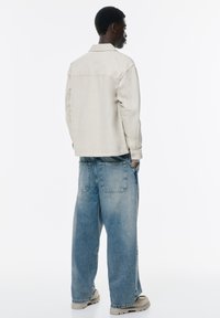 Beige denimjakke med krave og knappede manchetter, parret med oversized lyseblå jeans med store lommer. Neutralt tonet fodtøj.