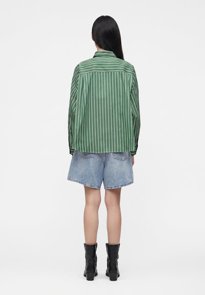 Personne aux longs cheveux foncés portant une chemise rayée verte et blanche, un short en jean bleu clair et des bottes noires mi-mollet, debout de dos.