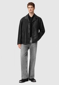 Νεαρός άνδρας φορέσει μαύρο δερμάτινο μπουφάν, μαύρο πουκάμισο, γκρίζα τζιν με ίσια γραμμή και μαύρα παπούτσια με λευκές σόλες, στέκεται σε έναν απλό φόντο.