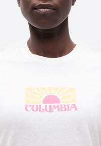 Κοντινό πλάνο ενός ατόμου που φοράει λευκό πουκάμισο με γραφικό ήλιο σε ροζ και κίτρινο χρώμα και κείμενο "COLUMBIA" στο στήθος, σε απλό φόντο.