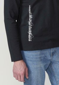 Hosszú ujjú fekete felső strukturált anyagból, fehér grafikus felirattal az oldalán. Világoskék farmerrel párosítva, ami egy laza megjelenést kölcsönöz.