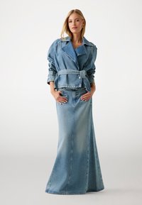 Giacca di denim con revers oversize e vita con cintura, abbinata a una lunga gonna di denim. Entrambi i pezzi sono di un blu chiaro con leggeri sbiadimenti.