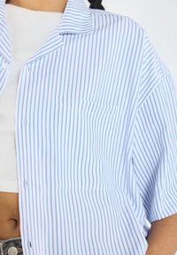 Camicia a righe verticali blu e bianche con maniche corte, colletto, taschino sul petto e una vestibilità ampia. Realizzata in un tessuto leggero.