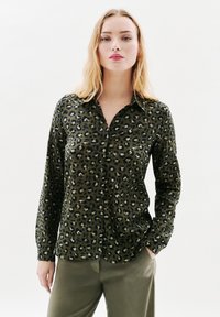 Giovane donna con lunghi capelli biondi che indossa una camicia a bottoni a stampa leopardata verde e nera e pantaloni verde oliva, in piedi davanti a uno sfondo bianco.