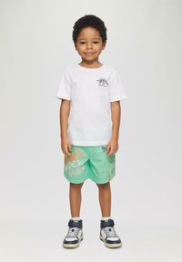 Weißes Baumwoll-T-Shirt mit einem Dinosaurier-Print, kombiniert mit grünen Shorts, die orangefarbene Schriftzüge und Spritzmuster aufweisen, getragen mit schwarzen und silbernen Turnschuhen.