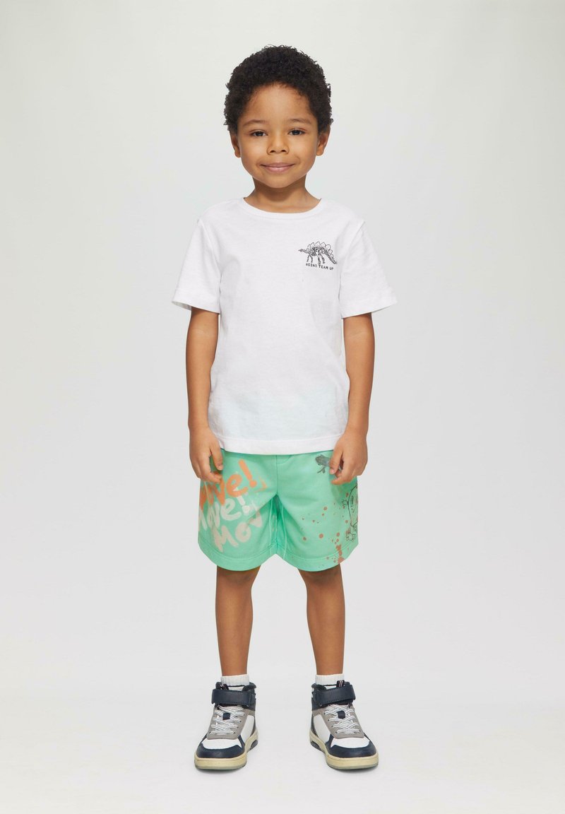 Weißes Baumwoll-T-Shirt mit einem Dinosaurier-Print, kombiniert mit grünen Shorts, die orangefarbene Schriftzüge und Spritzmuster aufweisen, getragen mit schwarzen und silbernen Turnschuhen.