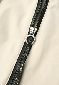 Gros plan sur une fermeture éclair noire avec un anneau en argent et une étiquette en tissu noir marquée "STREET ONE" sur un fond en tissu beige.