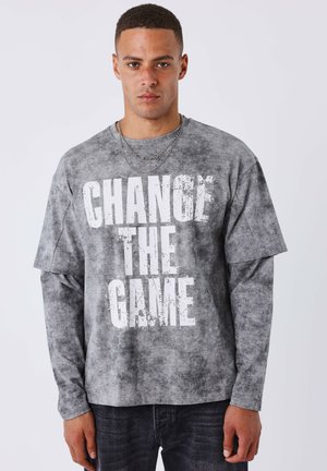 Giovane uomo con camicia grigia a strati con la scritta "CHANGE THE GAME" in lettere bianche in grassetto, in piedi davanti a uno sfondo chiaro e semplice.