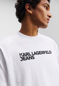 Weißes Baumwoll-T-Shirt mit einem großen schwarzen Logo, das "KARL LAGERFELD JEANS" zeigt. Einfaches Rundhalsdesign mit kurzen Ärmeln.