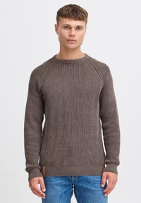 Brązowy sweter prążkowany z teksturowanym wzorem, okrągłym dekoltem i ściągaczami. Noszony z niebieskimi dżinsami, które mają małą metkę marki na dole.