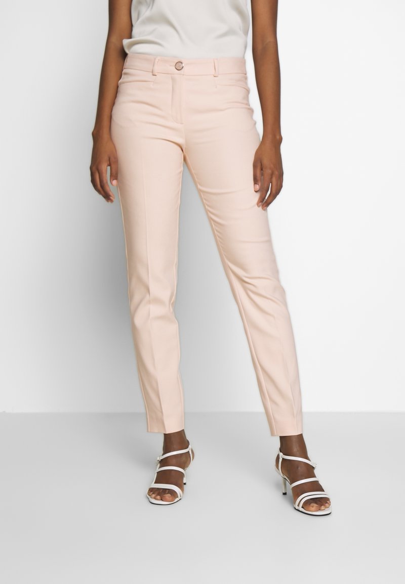 Femme portant un pantalon ajusté rose pâle et des sandales blanches à talons hauts et brides, debout devant un fond clair uni.