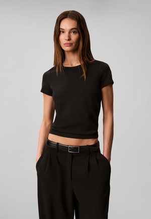 Ung kvinde med langt brunt hår iført en sort kortærmet crop top, sorte bukser og sort bælte med hænderne i lommerne.