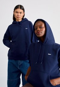 Dois modelos vestem hoodies azul-marinho com um bolso frontal e capuz ajustável. Cada um tem um logo branco "OBEY" no peito. O fundo é liso.