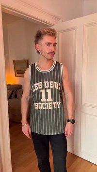 Giovane uomo con capelli chiari e baffi che indossa una maglietta a strisce senza maniche con la scritta "LES DEUX SOCIETY 11", in piedi in una porta interna.