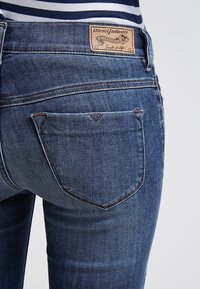 Mörkblå denimjeans med en texturerad yta, utrustad med en läderpatch med varumärket vid midjan och en trekantig bakfickdetalj.