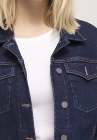 Mörk denimjacka med silverknappar, två framfickor och kontrasterande sömmar. Visad över en enkel vit t-shirt.