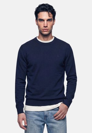 Uomo con capelli ricci scuri indossa un maglione blu navy sopra una camicia bianca e jeans azzurri, in piedi davanti a uno sfondo chiaro uniforme.