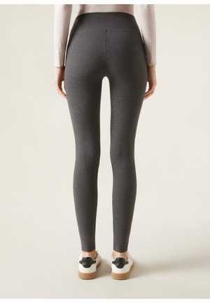 Høj talje, tætsiddende leggings i mørkegrå stof med en glat tekstur. Har en sømløs bagdel og bæres med hvide sko og sorte detaljer.