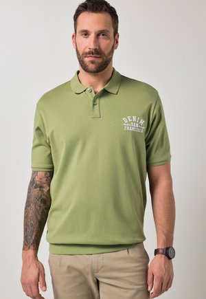 Mężczyzna z brodą i wytatuowanym ramieniem, ubrany w zieloną koszulkę polo z białym napisem "DENIM San Francisco" i beżowe spodnie, stojący na jasnym tle.