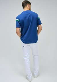 Camiseta de algodón azul con acentos a rayas verdes y blancas en las mangas, combinada con pantalones blancos y zapatillas gris claro. Modelo visto desde atrás.