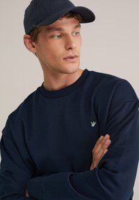 Temno modra majica z okrolim izrezom, z rebrastimi manšetami, ohlapnim krojem in majhnim vezenim logotipom na levi strani prsnega koša. Nosijo jo skupaj s primerno kapo.