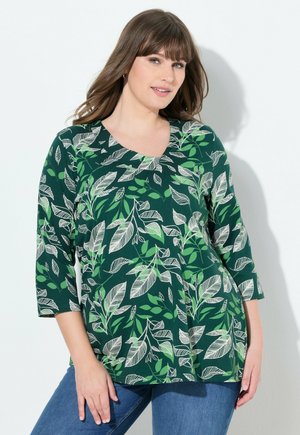 Mujer vistiendo una blusa verde con patrones de hojas blancas y verde claro, y jeans azules, posando contra un fondo blanco.