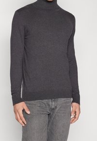 Mörkgrå polotröja i slät stickad kvalitetsmaterial; har långa ärmar och en figurnära design. Kombinerad med ljusgrå jeans.