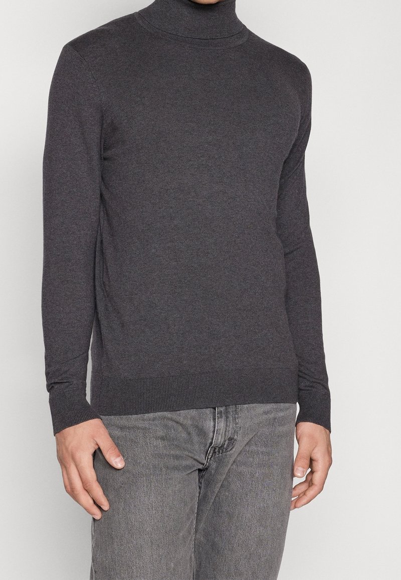 Mörkgrå polotröja i slät stickad kvalitetsmaterial; har långa ärmar och en figurnära design. Kombinerad med ljusgrå jeans.